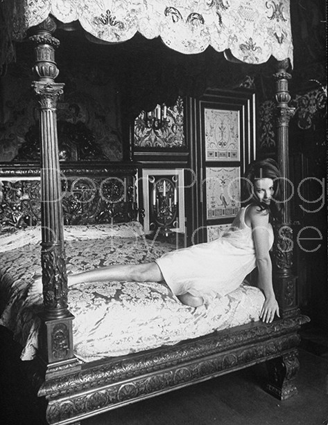 Actress Claudine Auger in lacy nightgown, leaning sensuously against canopy post of gorgeous hand-carved Regency bed from the Regency period in room at the Chateau d'Anet, the location for filming the movie "Thunderball".