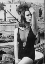 Actress Claudine Auger wearing a one-piece bathing suit that features a plunging fishnet neckline as she poses in front of fishing wharf backdrop.