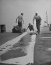 US naval personnel tarring cotton sheeting to the deck to preserve and seal the hull while the ship sits unused in a storage depot.