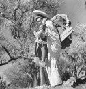 A man carrying a load of cut wood on his back to market, flowers strapped to his arm.
