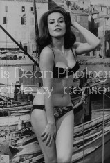 Actress Claudine Auger posing sexily in a bikini bathing suit in front of fishing wharf backdrop.