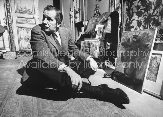 Actor Vincent Price sitting on the floor with his legs crossed in front of a few pieces of his art collection.